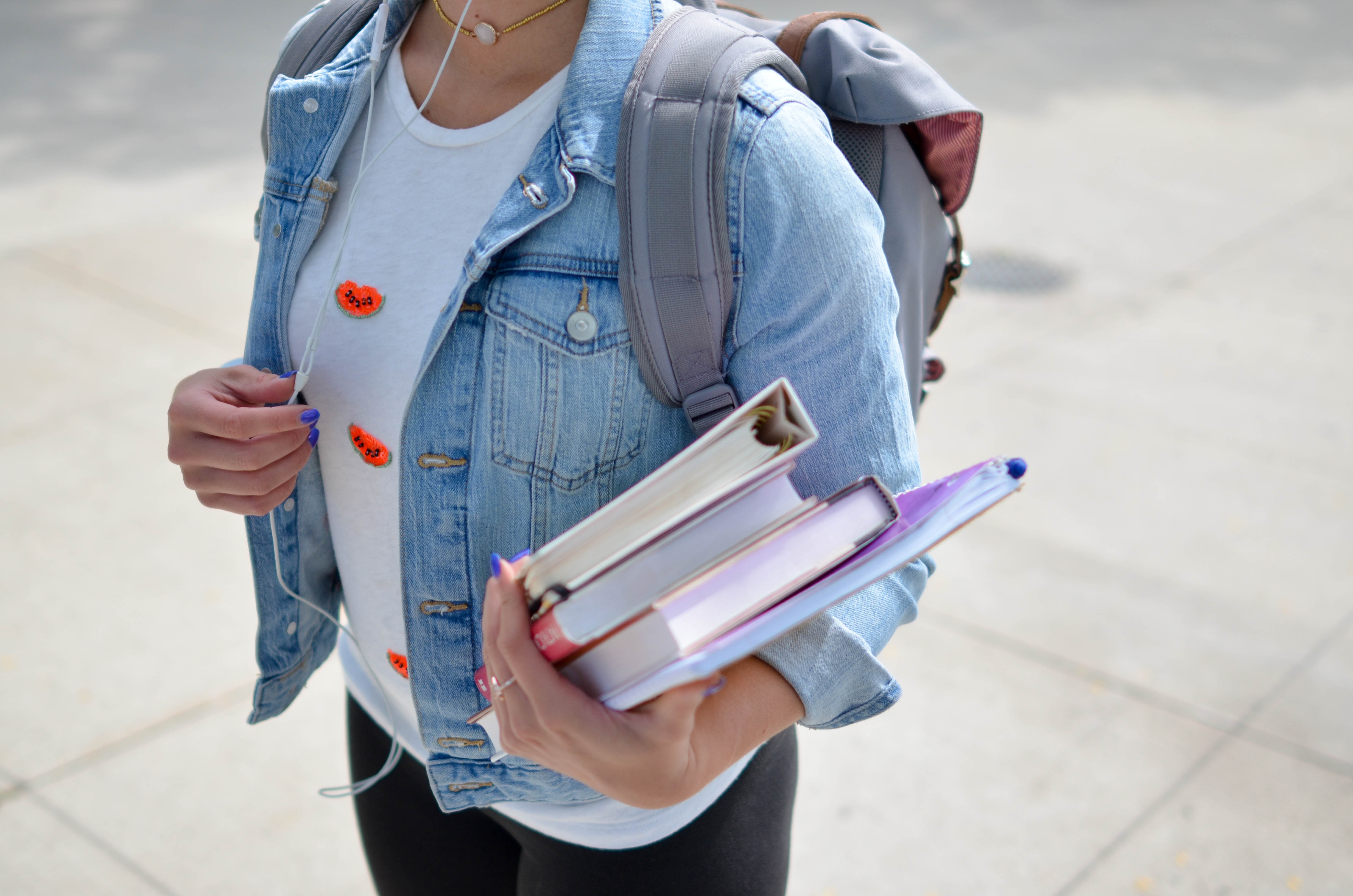 A girl wearing a white shirt and a jeans jacket, carrying some text books.